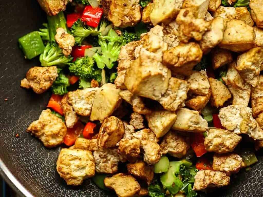 Stir-fried tofu cubes with broccoli, red bell peppers, and other vegetables in a frying pan. The tofu is golden brown, while the vegetables are vibrant and fresh.